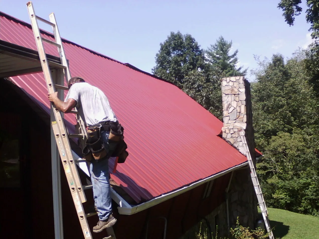 Roofer on a ladder beside a red metal roof and stone chimney — Soffit & Fascia Repair by Roofing Brothers in Belfield